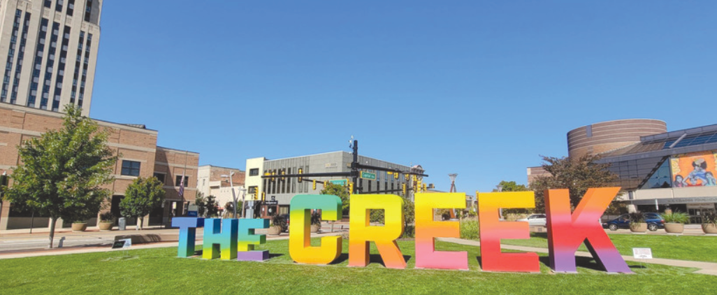 An art sculpture style, sign installation in the middle of a parkway near downtown Battle Creek, Michigan titled "The Creek" painted with all the colors of the rainbow blended into one long gradient.