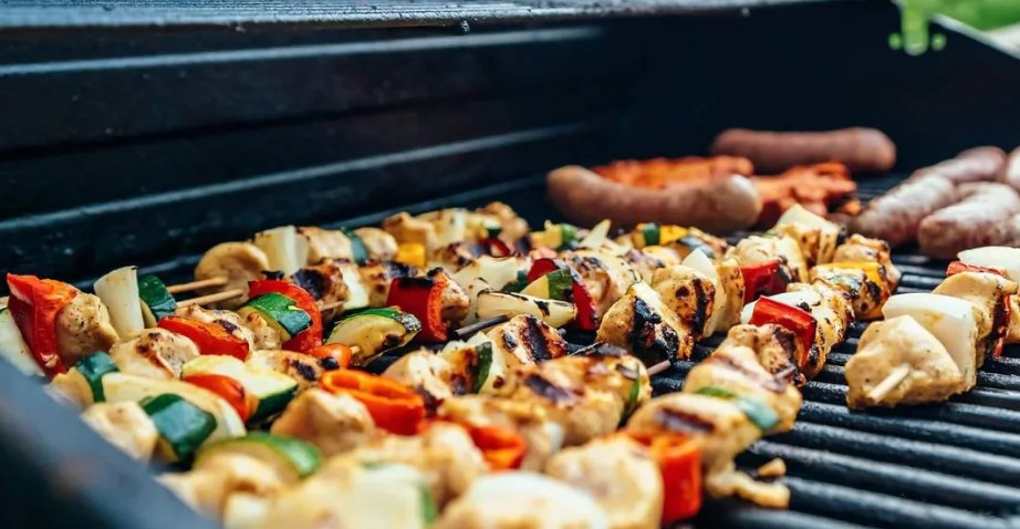 A photo of an open barbeque grill with a several sizzling chicken and vegetable kabobs, sausages, and other all-a-cart vegetables.