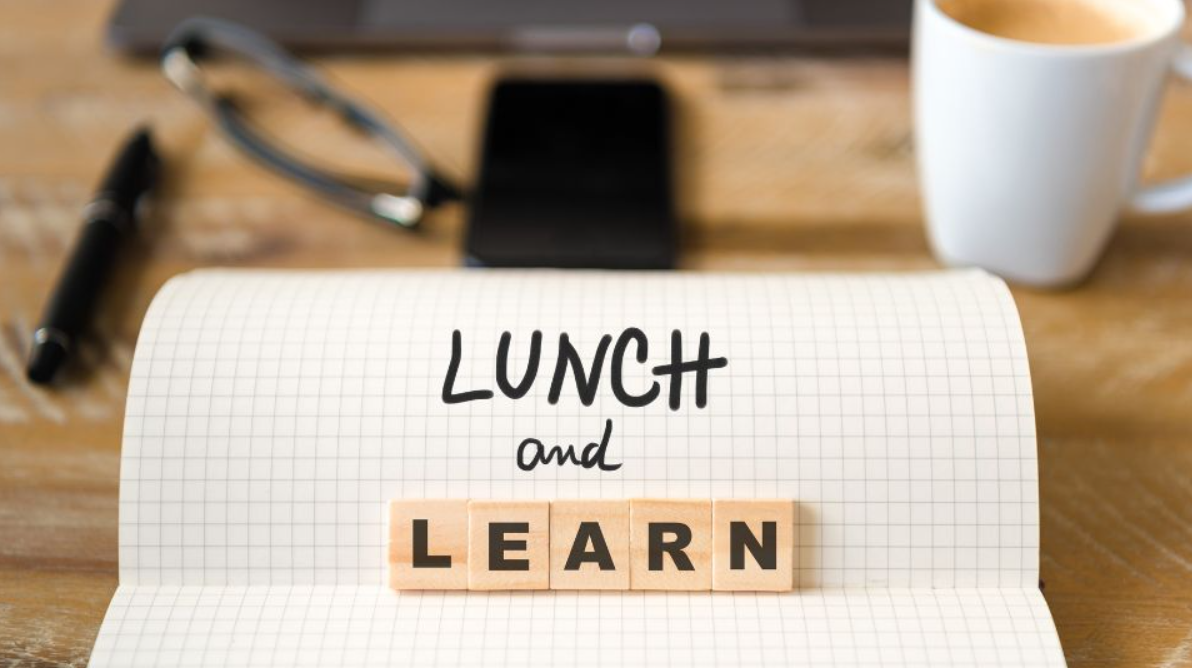 An office desk with coffee mug, eyeglasses, and pen among a rolled sheet of graph lined paper and the words "Lunch and Learn" where L.E.A.R.N. are scrabble pieces under the words Lunch and that have been written out.