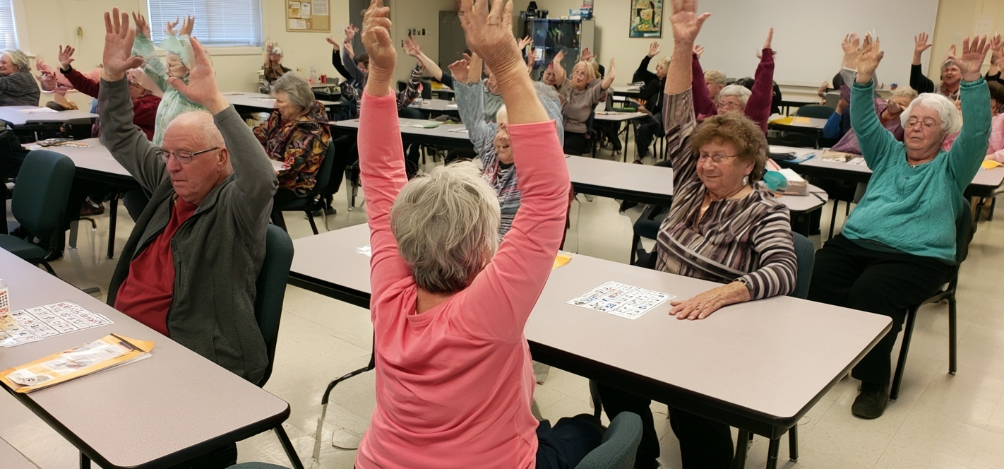 A group of seniors in a classroom setting, seated and tables while playing Bingo and performing light chair exercises.