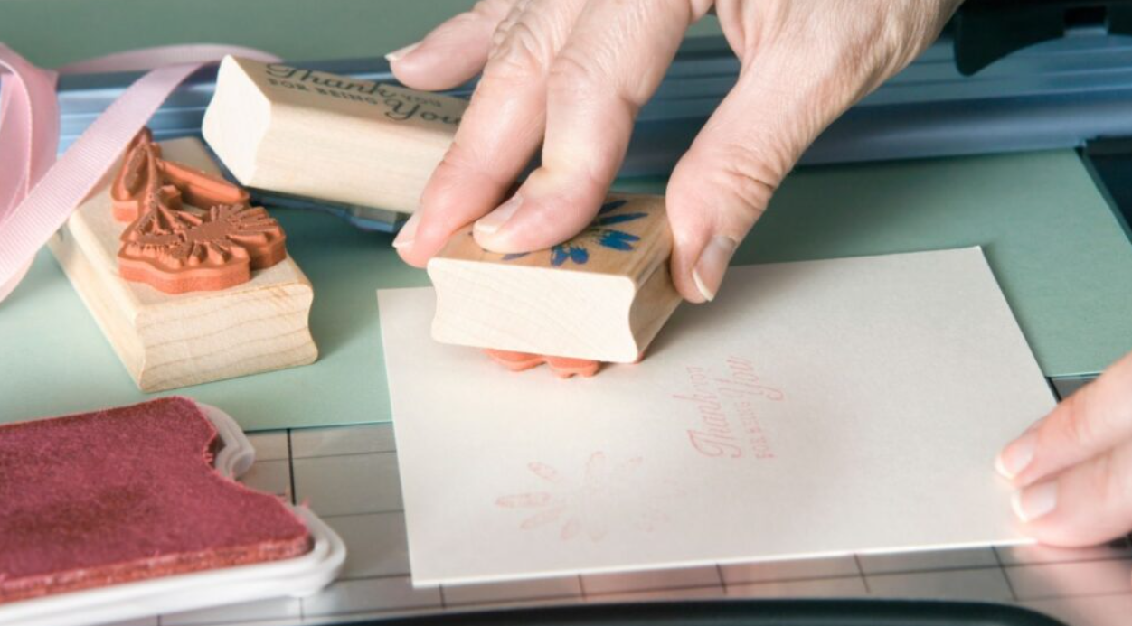 A set of hands pressing down on a rubber ink stamp used for paper crafts with other inking materials around the table.