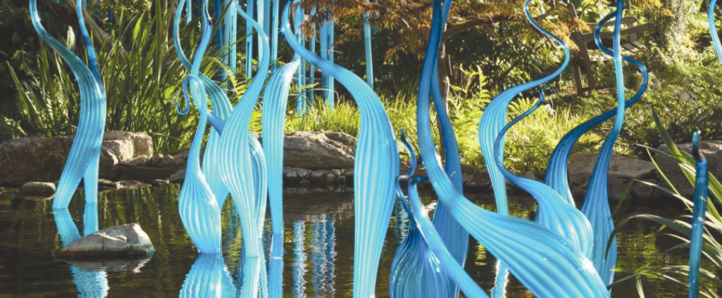 An image depicting an art installation of world-renowned artist Dale Chihuly. The glass artwork resembles sea plants, similar to the blades of kelp seaweed and other snake-like plants emerging from a water garden feature from inside the Frederik Meijer Gardens and Sculpture Park.