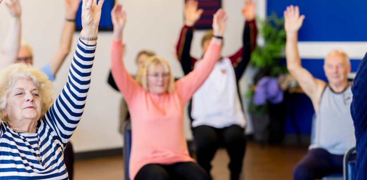 A group of people sitting in chairs with their arms raised high as part of an exorcise routine.