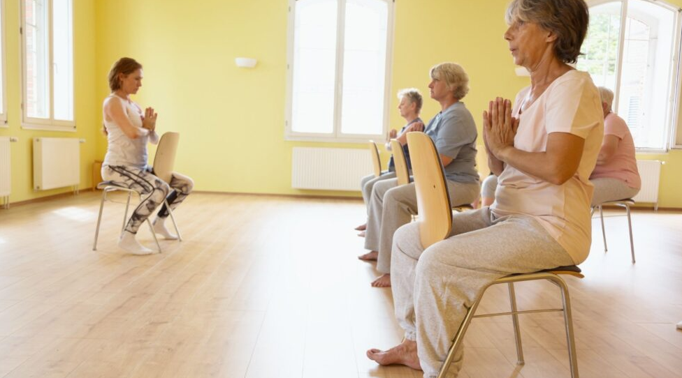 A group of participants in a classroom setting seated on chairs doing modified simple yoga techniques with an instructor.