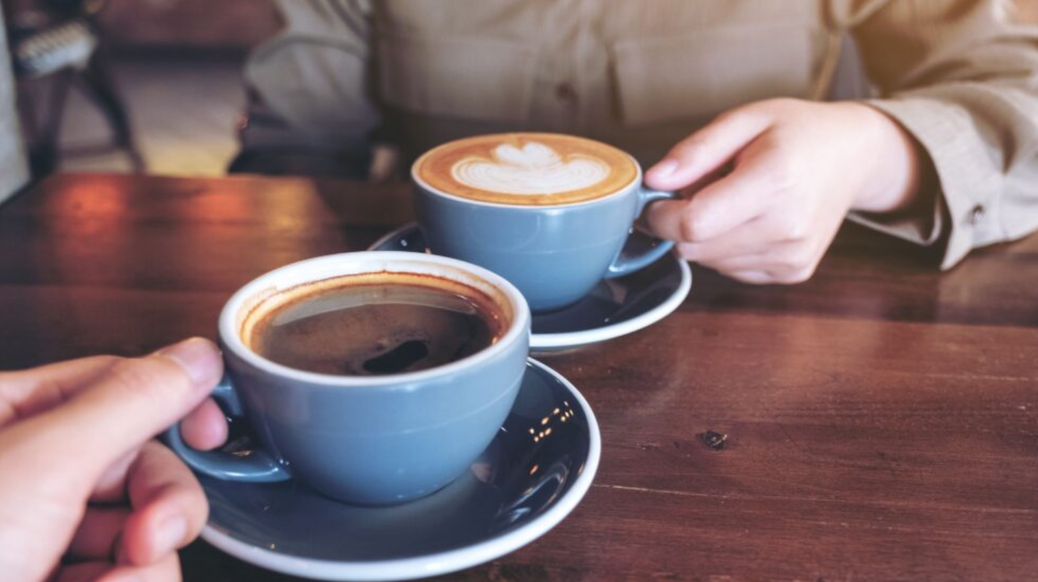 Two friends sitting at a café table with their hands holding coffee while engaging in conversation.