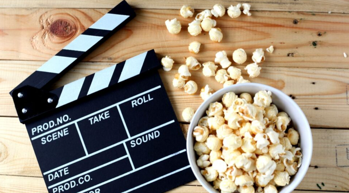 An image of a film clapper next to a bowl of fresh popcorn set upon a wood table.