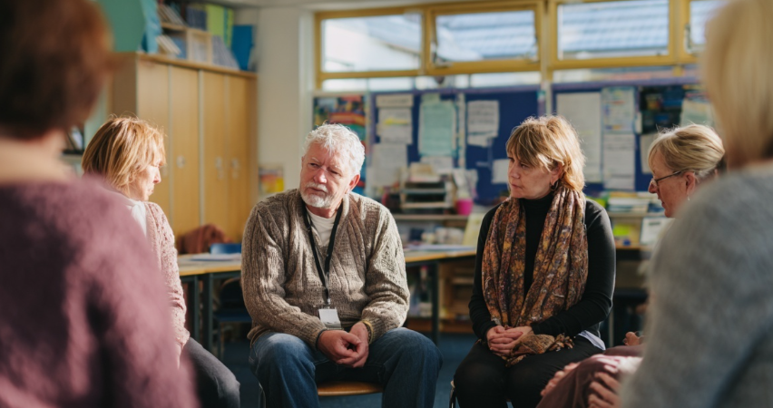 A group of members seated in a support group style setting sharing stories of comfort and grief.