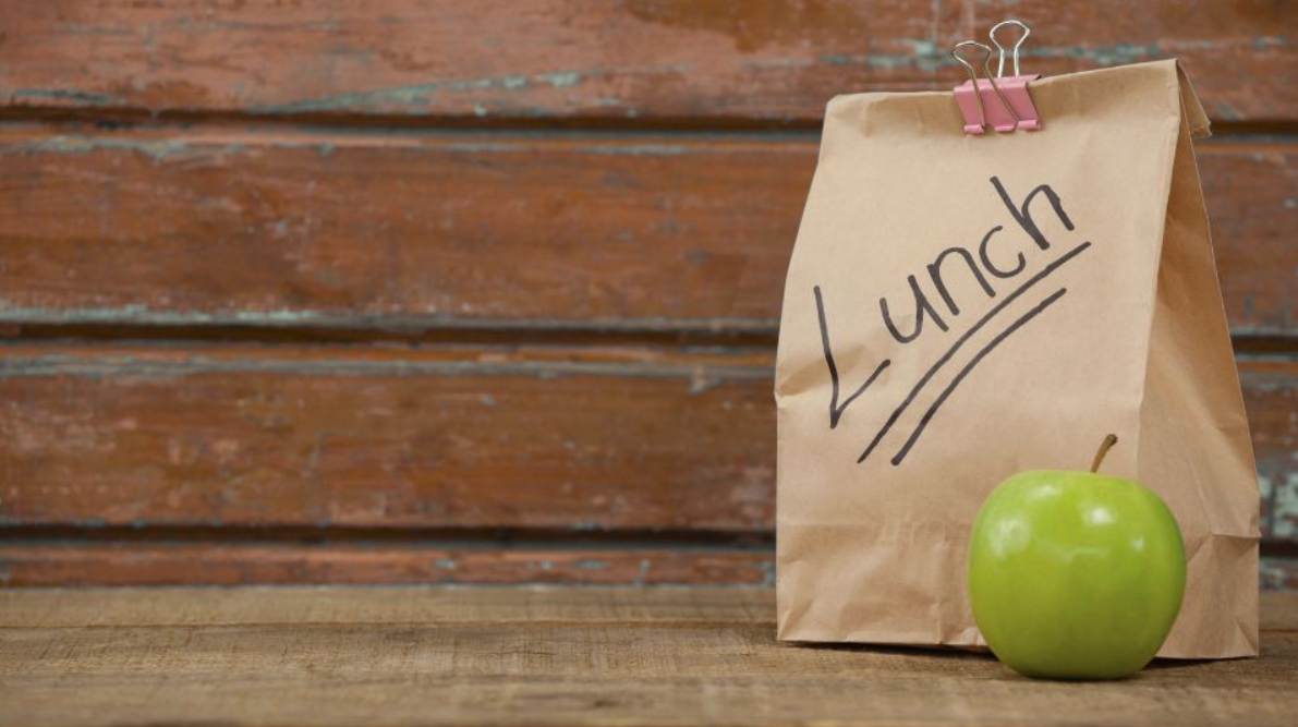 A brown paper bag with the word "Lunch" underlined. The bag is closed with a office binder clip and sits on a table next to an apple.