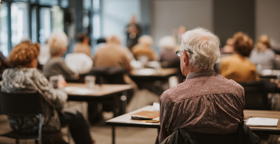 A group of members in a classroom setting focused on a speaker at the front of the room while having snacks in a Lunch & Learn opportunity.