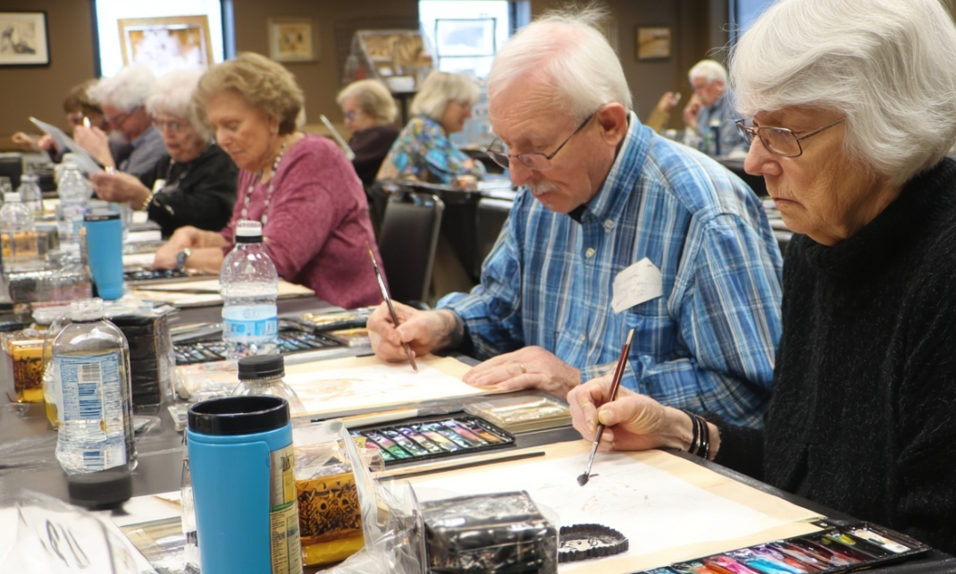 A group of members sitting at table in a classroom-like setting working on individual paintings.