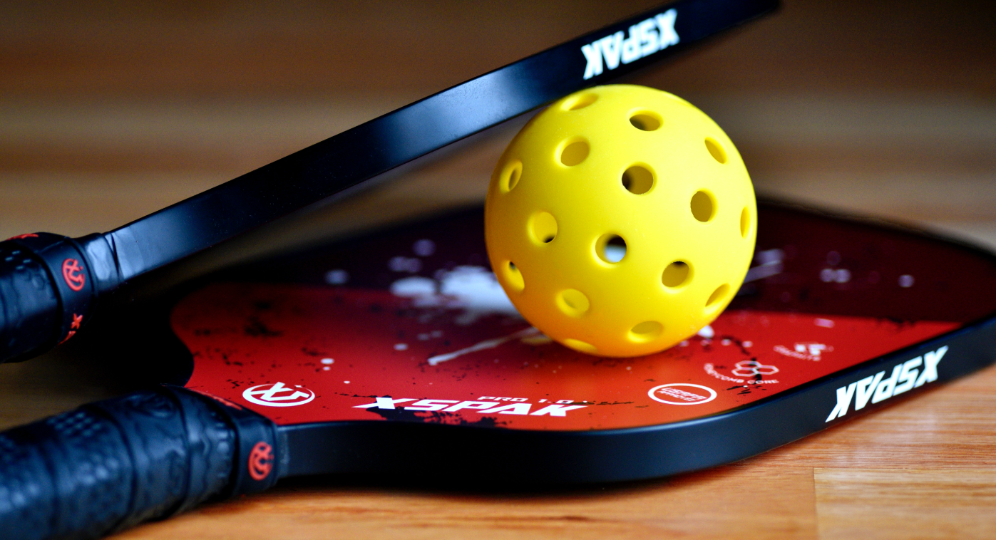 A couple of pickleball paddles and a ball resting on a gymnasium floor.