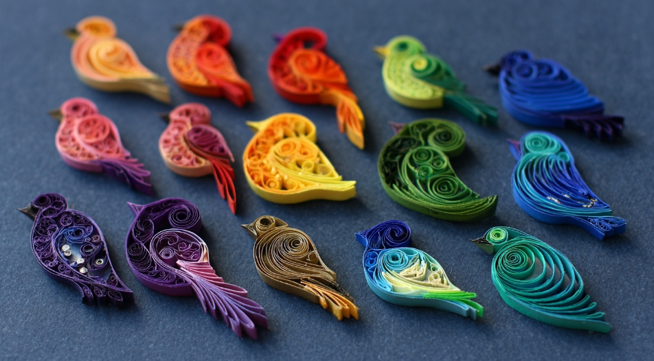 A display of quilled birds of all shapes and colors made from rolled paper sitting in alignment on a table.