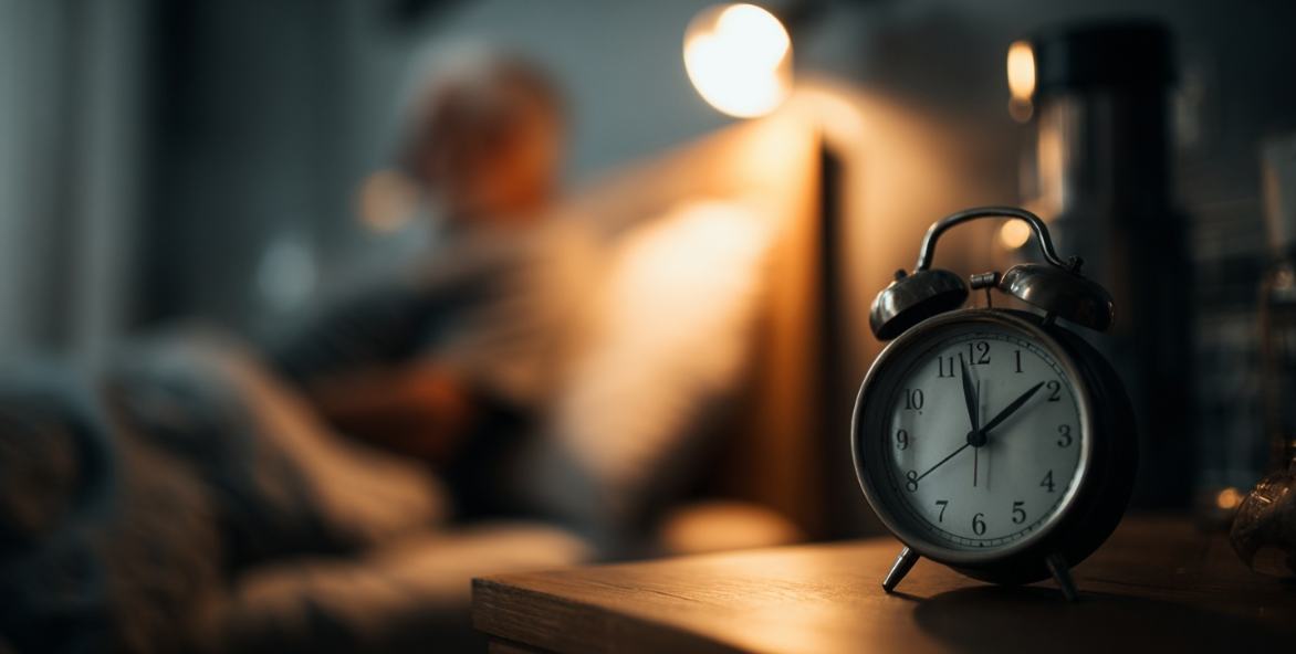 A photo of an alarm clock on a nightstand, late at night with someone laying in bed with a nightlight turned on, getting ready to sleep for the night.