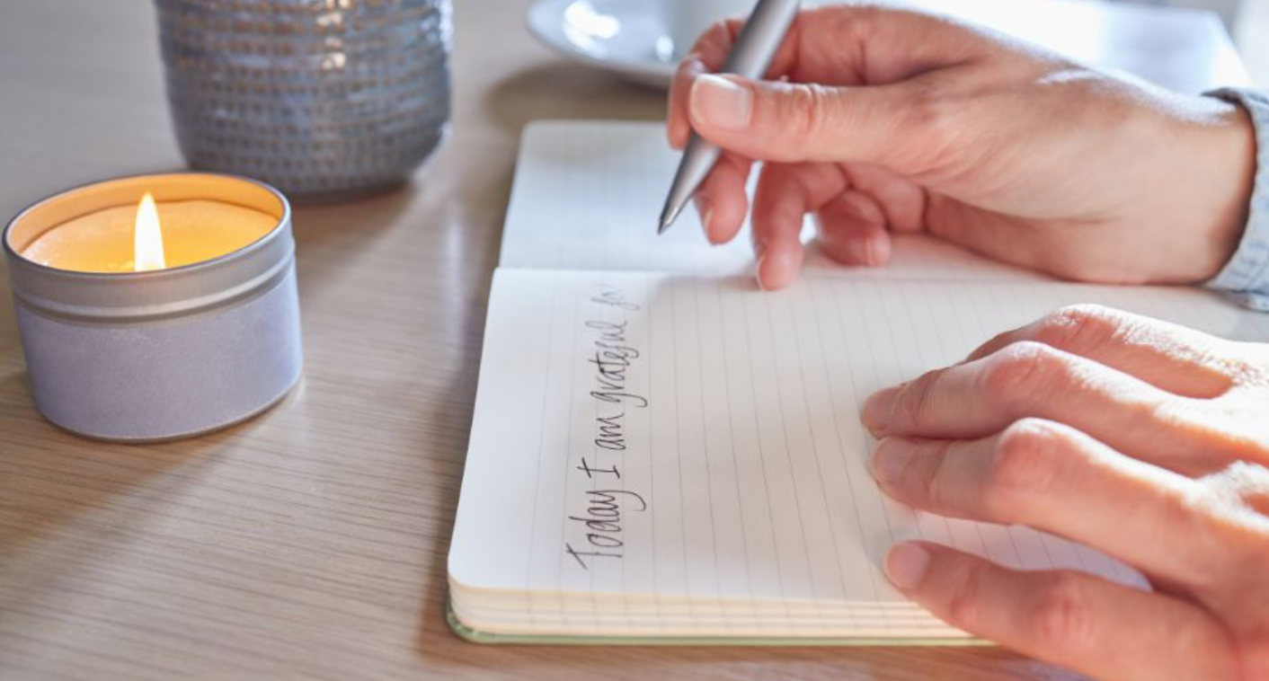 A person sitting at a writing desk with a notebook open and a pen in their hand, writing text between the lines.